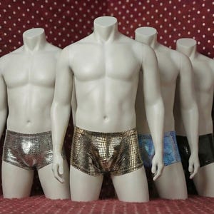 以下が含まれることがあります： 銀、金、青、黒のメタリック柄ボクサーパンツを展示する4体の頭のないマネキン。マネキンは白い水玉模様の入ったボルドー色の背景に立ち、下着の質感と色を際立たせています。