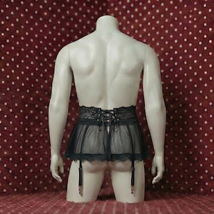 Könnte beinhalten: Ein schwarzer Dessous-Rock mit Spitzendetails und Strumpfhaltern wird auf einer Schaufensterpuppe präsentiert. Der Rock besteht aus durchsichtigem Stoff und hat einen Schnürrücken. Die Schaufensterpuppe steht vor einem roten Hintergrund mit weißen Punkten.