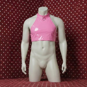Puede incluir: Top halter rosa brillante con detalle de anillo en el cuello, exhibido en un maniquí. El top es de estilo crop top. El maniquí está sobre un fondo rojo con lunares blancos.
