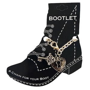 Pode incluir: Uma corrente de bota prateada com um pingente de chave e cadeado em forma de coração, exibida em um cartão preto em forma de bota. O cartão diz "BOOTLET" e "A CHAIN FOR YOUR BOOT."