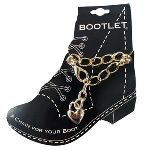 Pode incluir: Corrente de bota dourada com um pingente em forma de coração. A corrente é exibida em um cartão preto em forma de bota, com o texto "BOOTLET" e "A CHAIN FOR YOUR BOOT".