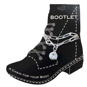 Pode incluir: Uma corrente de bota prateada com um pingente iridescente pendurado, exibida em um cartão preto em forma de bota. O cartão diz "BOOTLET" e "A CHAIN FOR YOUR BOOT."