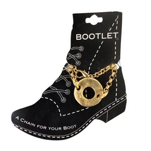 Pode incluir: Um cartão preto em forma de bota com a palavra "BOOTLET" no topo. Uma corrente dourada com um pingente circular está presa. O texto "A CHAIN FOR YOUR BOOT" está na parte inferior.