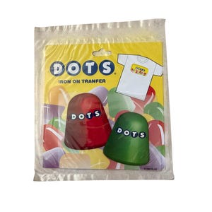 Könnte beinhalten: Eine Packung Dots-Bügelbilder. Die gelbe Rückkarte zeigt das Wort "DOTS" in Blau, eine weiße T-Shirt-Grafik und zwei bonbonförmige Bügelbilder, eines rot und eines grün, jeweils mit "DOTS" aufgedruckt. Die Verpackung ist in klarem Kunststoff versiegelt.