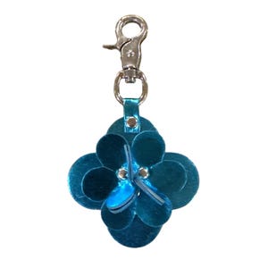 May include: A teal blue leather keychain with a flower design. The flower has four rounded petals and a smaller, layered flower in the center. A silver-colored clasp and a small leather loop connect the flower to the keychain.