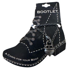 Pode incluir: Uma etiqueta preta em forma de bota com a palavra "BOOTLET" no topo. Uma corrente com um pingente decorativo pende da etiqueta. O texto "A CHAIN FOR YOUR BOOT" está impresso na etiqueta.