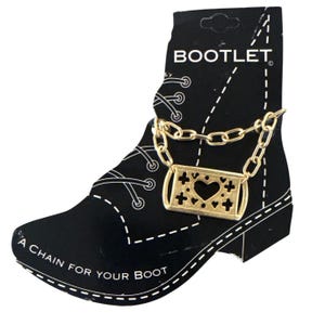Op de afbeelding: Een goudkleurige laarketting met een rechthoekige hanger met een hartuitsparing en kruisontwerpen. De ketting wordt getoond op een zwarte kaart in de vorm van een laars, met de tekst "BOOTLET" en "A CHAIN FOR YOUR BOOT".