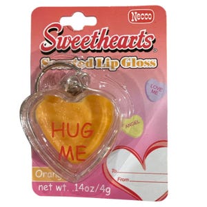 May include: Orange heart-shaped lip gloss with the words "HUG ME" in red, packaged on a pink card. The card features the "Sweethearts" logo and the text "Scented Lip Gloss". A small heart-shaped candy with "LOVE ME" is also visible.