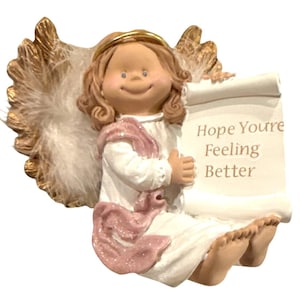 Puede incluir: Figura de ángel de cerámica con aureola dorada y alas emplumadas. El ángel viste una túnica blanca y una faja rosa, sosteniendo un pergamino que dice "Hope You're Feeling Better."