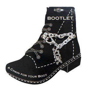 Pode incluir: Um acessório de bota de corrente prateada com fecho lagosta e berloques decorativos, exibido num cartão preto em forma de bota. O cartão diz "BOOTLET" e "A CHAIN FOR YOUR BOOT" em texto branco.