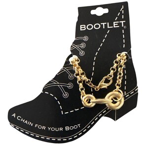 Pode incluir: Corrente de bota dourada com fecho e design decorativo de freio. A corrente é exibida em um cartão preto em forma de bota, com o texto "BOOTLET" e "A CHAIN FOR YOUR BOOT".