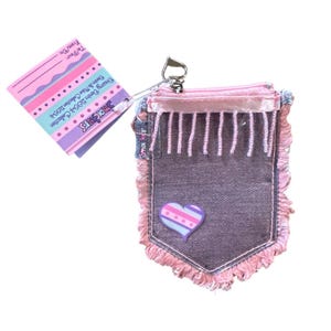 Könnte beinhalten: Eine kleine Geldbörse im Jeans-Stil mit rosa Reißverschluss und Fransen. Die Geldbörse hat einen herzförmigen Aufnäher mit Streifen und Punkten. Ein Etikett trägt die Aufschrift "Dreamy Denim 2004 Collection" und "Smackers."