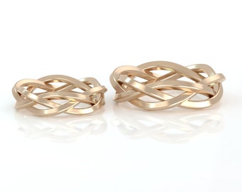 Puede incluir: Dos anillos trenzados de oro, uno más grande que el otro, se muestran sobre un fondo blanco. Los anillos están hechos de una sola pieza de metal que se entrelaza para crear un diseño complejo.