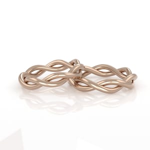May include: Two intertwined gold wedding bands, each with a braided design, are displayed on a white background.