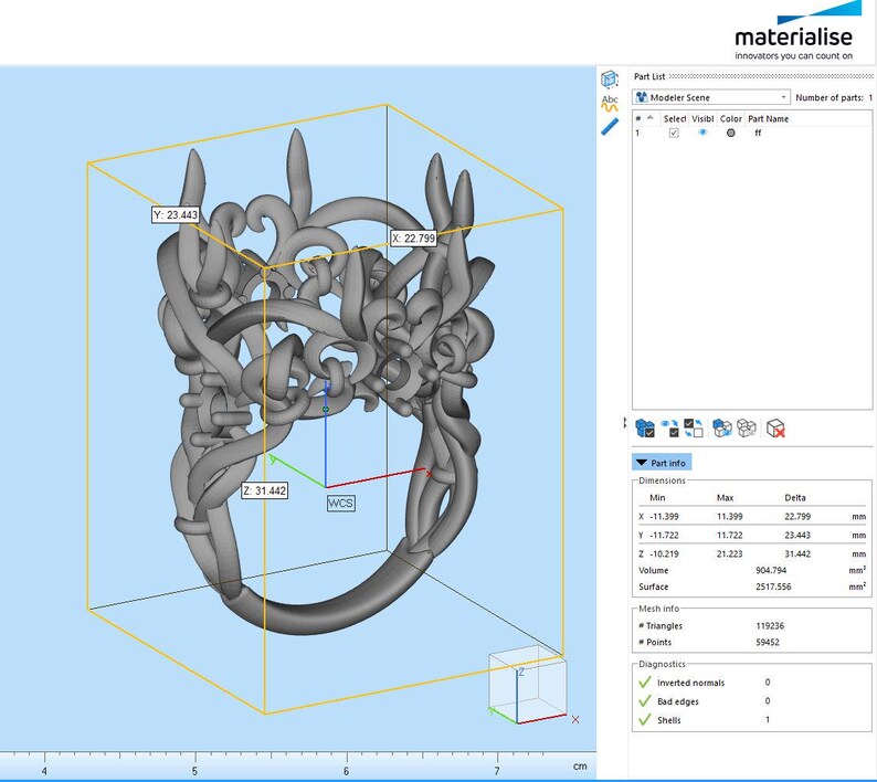 Puede incluir: Un modelo digital 3D gris de un anillo con un dise&ntilde;o org&aacute;nico intrincado similar a enredaderas, mostrado en el software Materialise. La interfaz muestra dimensiones de 22,799 mm por 23,443 mm por 31,442 mm en un cuadro amarillo sobre fondo azul.