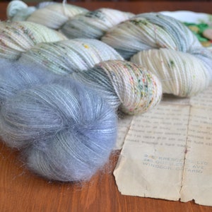 May include: Two skeins of yarn in a light blue and white colourway with speckles of green and brown. The yarn is arranged in a loose coil. A third skein of yarn in a light blue colourway is in the foreground.