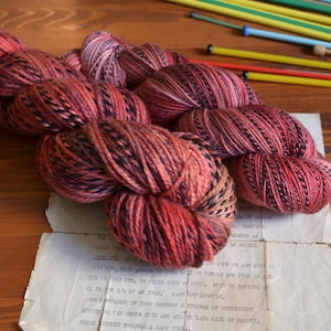 May include: Two skeins of yarn in a deep red and black colourway. The yarn is twisted together and has a speckled appearance.