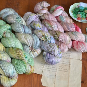 Könnte beinhalten: Sechs Knäuel buntes Garn in Grün-, Rosa- und Lilatönen. Das Garn ist in einem Regenbogenmuster angeordnet. Das Bild zeigt auch Stricknadeln und Knöpfe.