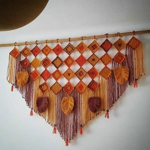 Op de afbeelding: Een gehaakte wandhanger met een geometrisch patroon in tinten oranje, bruin en geel. De hanger heeft een franje rand en bladvormige accenten.