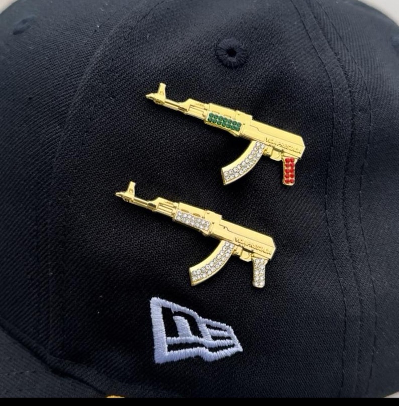 Puede incluir: Dos pines AK-47 dorados con detalles de pedrer&iacute;a y detalles de color, exhibidos en una gorra de b&eacute;isbol negra. Los pines tienen detalles verdes y rojos. La gorra presenta un logotipo blanco.