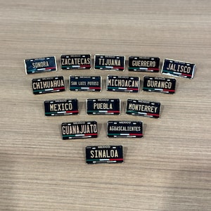 May include: A collection of 14 black and gold Mexican state license plate pins. Each pin features the name of a Mexican state in gold lettering on a black background with a red, white, and green Mexican flag.