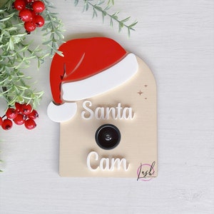 May include: A wooden sign with a Santa hat on top. The sign says "Santa Cam" and has a black camera lens in the middle.
