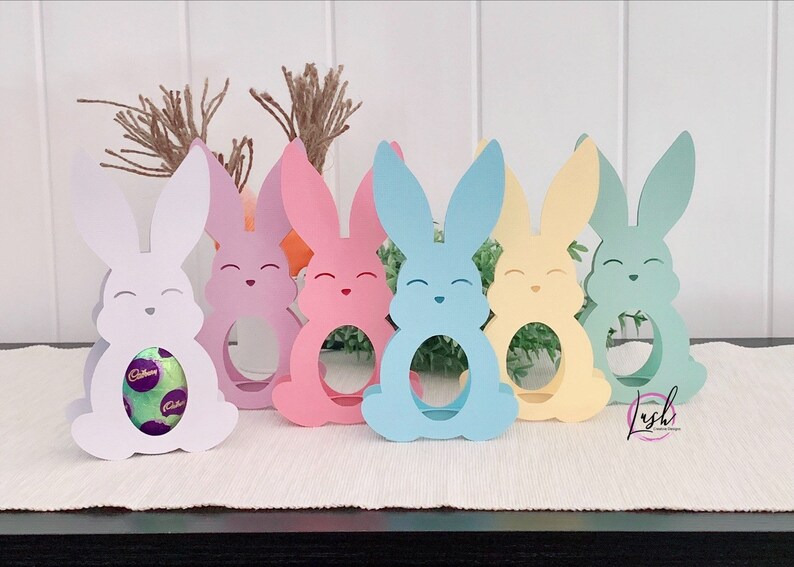 May include: A collection of colorful, laser-cut Easter bunny-shaped holders. Each bunny has a smiling face and a circular opening to hold a treat. The bunnies are in shades of white, pink, blue, yellow, and green, perfect for Easter decorations.