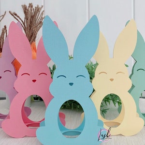 May include: A collection of pastel-colored paper bunny cutouts. The bunnies are in shades of pink, blue, yellow, and green, each with a circular opening in the center. They have smiling faces and are likely Easter decorations.