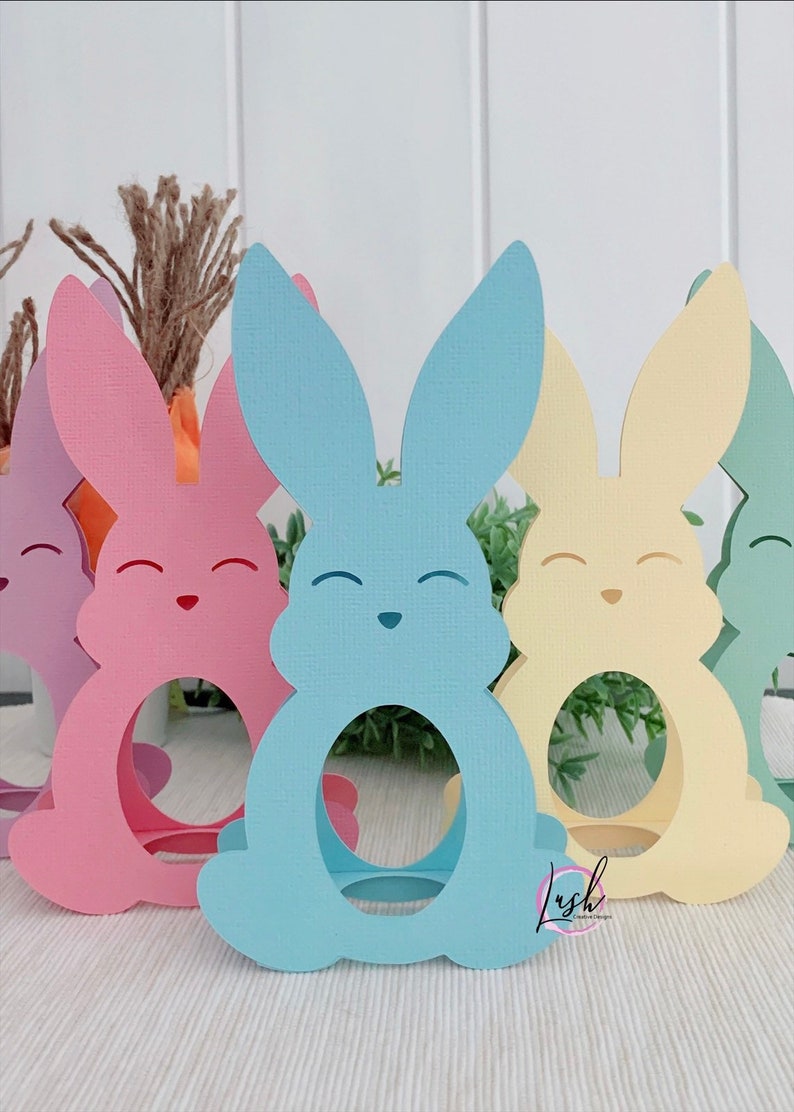 May include: A collection of pastel-colored paper bunny cutouts. The bunnies are in shades of pink, blue, yellow, and green, each with a circular opening in the center. They have smiling faces and are likely Easter decorations.