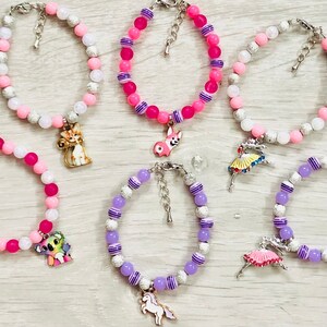 Peut inclure: Cinq bracelets en perles colorées avec des breloques. Les bracelets sont faits de perles roses, blanches et violettes. Les breloques comprennent un chat, un chien, une ballerine, un koala et une licorne.