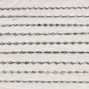 May include: A collection of 10 silver chains with different link styles, including round, heart, cross, and diamond shapes. The chains are laid out horizontally on a white wooden surface.