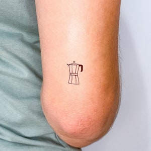 Puede incluir: Un dibujo de línea simple de una cafetera, o cafetera Moka, en el brazo de una persona. El dibujo está hecho con tinta negra y es un diseño de tatuaje popular.