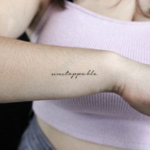 Op de afbeelding: Een close-up van een arm met een kleine, zwarte tatoeage. De tatoeage luidt "unstoppable" in een sierlijke, cursieve lettertype. De huid is licht, en de arm is licht gebogen, met een lichtroze top op de achtergrond.