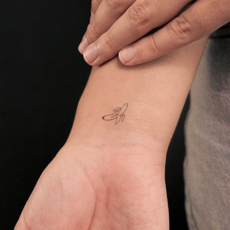 Pode incluir: Uma pequena tatuagem minimalista de uma banana descascada no pulso. A tatuagem &eacute; um contorno simples em tinta preta, representando a forma e os detalhes da banana. O tom de pele &eacute; claro e o fundo &eacute; escuro.