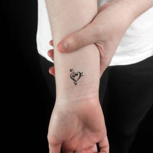 Op de afbeelding: Een kleine, zwarte tatoeage op een pols met een muzikale G-sleutel en twee stippen die een hartvorm vormen. De tatoeage bevindt zich op een blanke pols, zachtjes vastgehouden door een hand. De achtergrond is donker.