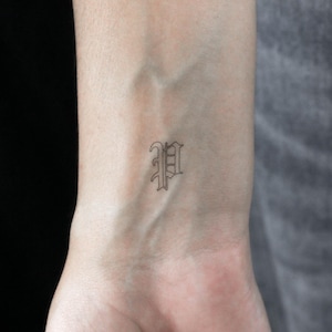 Può includere: Un primo piano di un polso con un piccolo tatuaggio della lettera "P" in un font gotico.