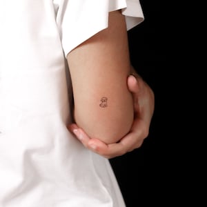 Könnte beinhalten: Ein kleines, minimalistisches Tattoo in schwarzer Tinte, das einen sitzenden Welpen mit Schlappohren und einem einfachen Umrissdesign zeigt. Das Tattoo befindet sich auf der Haut und zeichnet sich durch klare Linien und eine einfache Strichzeichnung aus.