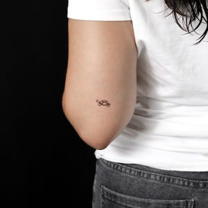 May include: A small, minimalist tattoo of a turtle, rendered in black ink. The turtle is depicted in a simple, line-art style, with a rounded shell and visible head and legs. The tattoo is located on the inner arm, near the elbow.