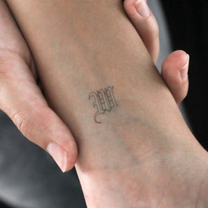 Puede incluir: Primer plano de un tobillo con un pequeño tatuaje de la letra "W" en fuente gótica.