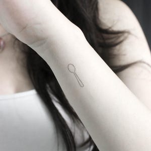 May include: A simple line drawing tattoo of a spoon on a person's wrist.