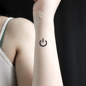 Può includere: Un primo piano di un polso con un piccolo tatuaggio nero a forma di pulsante di accensione.