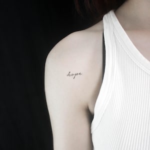 Könnte beinhalten: Nahaufnahme einer Schulter mit einem kleinen Tattoo, auf dem in Schreibschrift "Hoffnung" steht.