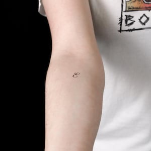 Puede incluir: Un pequeño tatuaje temporal negro de la letra E en cursiva sobre la piel de un brazo. El diseño presenta una fuente de escritura fluida y delicada con un bucle elegante, colocado en el centro del antebrazo sobre un fondo liso.