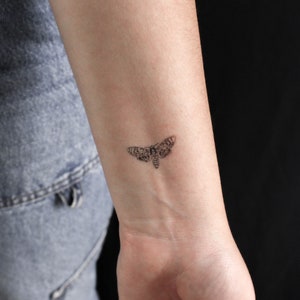 Könnte beinhalten: Ein kleines schwarz-weißes Motten-Tattoo auf dem Handgelenk einer Person.