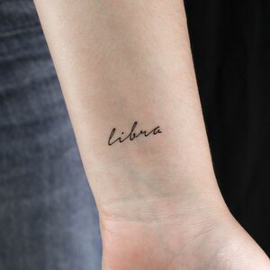 May include: A close-up of a person's wrist with a small tattoo that says "libra" in cursive script.