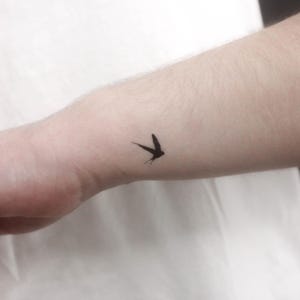 May include: A small, black swallow tattoo on the wrist. The bird is depicted in flight, with its wings spread. The tattoo is a simple, minimalist design, and the black ink contrasts with the skin tone.
