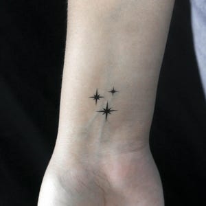 May include: A simple tattoo design of three small, black, starburst-shaped stars on a person's wrist.
