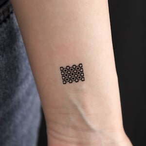 May include: A small black geometric tattoo design on a person's wrist. The design is made up of a series of squares arranged in a grid pattern.