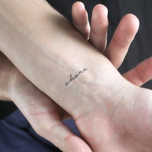 Könnte beinhalten: Nahaufnahme eines Handgelenks mit einem kleinen Tattoo. Das Wort "ahora" ist in Schreibschrift mit schwarzer Tinte geschrieben. Die Haut ist hell, und das Tattoo befindet sich am inneren Handgelenk.