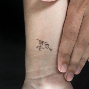 Könnte beinhalten: Ein kleines, schwarzes Tattoo in Linienzeichnung eines Zentauren mit einem Speer. Der Zentaur ist in einer laufenden Pose dargestellt, mit hochgehaltenem Speer. Das Tattoo befindet sich auf einem Handgelenk, der Hautton ist sichtbar.